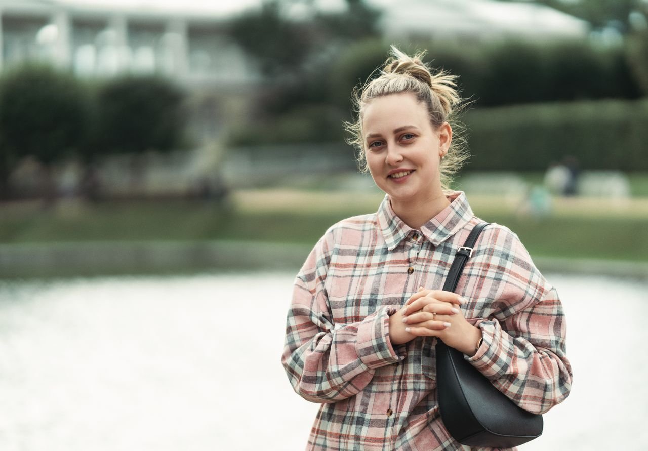 A woman in casual attire standing by a lake smiling outdoors, conveying a relaxed mood.